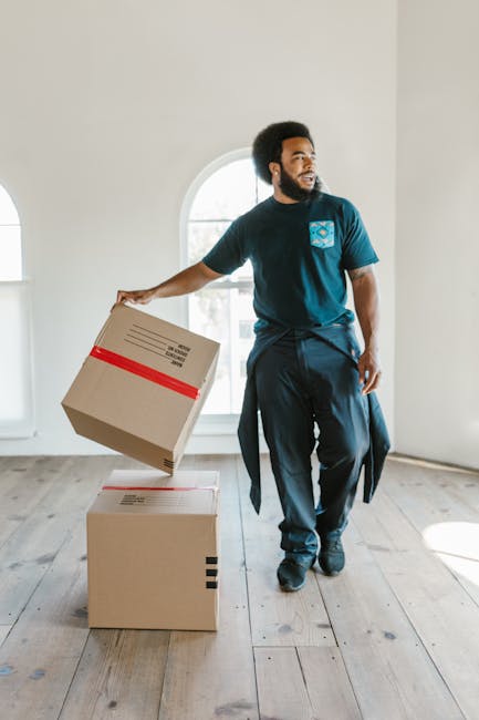 A man with dark curly hair and a beard, dressed in a dark blue t-shirt and trousers, is inside a well-lit room with white walls and wooden flooring during a home relocation. He is holding a cardboard box with red tape, preparing to place it on another similar box positioned on the floor. Behind him, a large arched window allows natural light to illuminate the space. The room appears to be empty, suggesting a moving process in progress, with the man part of a professional removals service. The scene captures the careful packing and handling involved in furniture transport and packing during a house move, with potential inclusion of moving blankets or trolleys used elsewhere in the process. This image exemplifies the logistical aspect of house removals, aligning with services offered by companies like Man With a Van King's Cross, operating within the removals sector, and highlights aspects of loading and packing for a successful move.