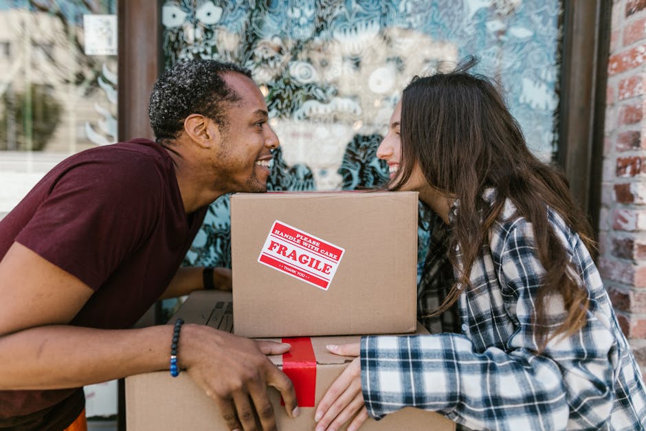 A man and a woman are positioned face-to-face outside a building, smiling at each other while holding or exchanging cardboard boxes used for home relocation. The man, with curly hair and wearing a maroon t-shirt and a black bracelet, is leaning in slightly towards the woman. The woman, with long brown hair and a plaid shirt, is also leaning forward, holding a cardboard box with red tape and a red and white 'fragile' handling sticker visible on the top box. The background features a large window with decorative frosted patterns and a brick wall. They are engaged in the packing and moving process, reflecting the activities related to furniture transport and house removals. The scene appears to be on a pavement or outdoor loading area, with natural daylight illuminating the scene, emphasizing the professionalism of a removals service like Man With a Van King's Cross involved in packing, loading, and furniture transport activities.