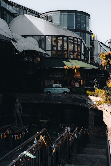 Exterior view of a modern multi-storey building with large glass windows and curved architectural elements, situated above a retail or dining area with visible signage for 'Cameo Market.' The building features white and black facades, with some outdoor lighting and potted plants contributing to its contemporary appearance. A white van with a visible logo is parked underneath on a cobbled street, indicating transportation or moving activity. In the foreground, metal railings and steps lead down to a lower level, where a person is walking nearby. The environment appears to be in an urban setting, with natural daylight illuminating the scene, suggesting daytime. This image reflects the context of home relocation or furniture transport, highlighting the logistics involved in moving furniture into or out of commercial or residential buildings, as handled by professional removals services such as Man With a Van King's Cross.
