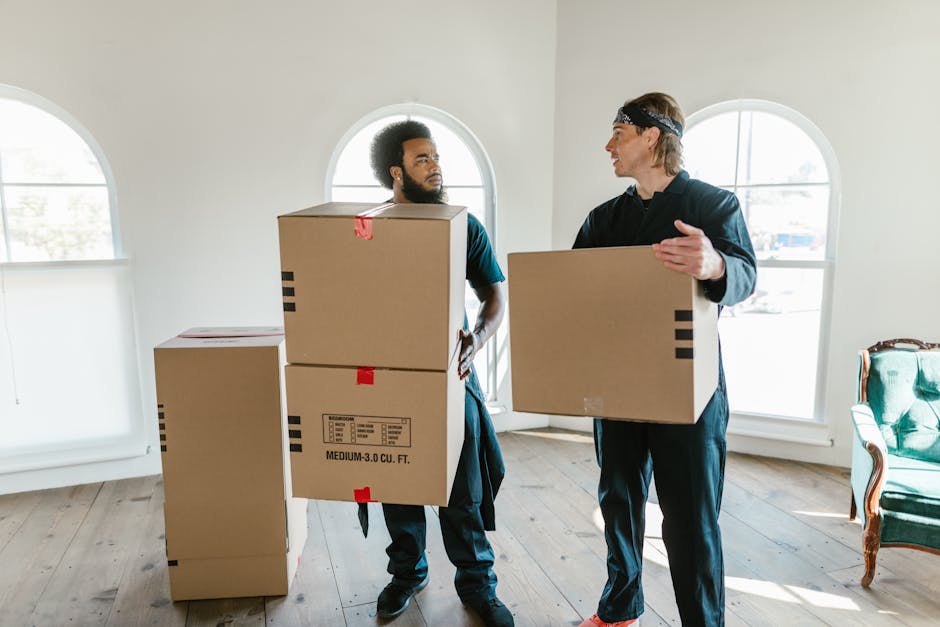 A man and a woman are positioned face-to-face outside a building, smiling at each other while holding or exchanging cardboard boxes used for home relocation. The man, with curly hair and wearing a maroon t-shirt and a black bracelet, is leaning in slightly towards the woman. The woman, with long brown hair and a plaid shirt, is also leaning forward, holding a cardboard box with red tape and a red and white 'fragile' handling sticker visible on the top box. The background features a large window with decorative frosted patterns and a brick wall. They are engaged in the packing and moving process, reflecting the activities related to furniture transport and house removals. The scene appears to be on a pavement or outdoor loading area, with natural daylight illuminating the scene, emphasizing the professionalism of a removals service like Man With a Van King's Cross involved in packing, loading, and furniture transport activities.