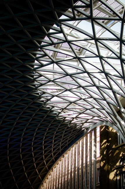 A detailed view of a modern architectural structure featuring a complex network of metallic beams and girders forming a curved, lattice-like roof. The framework consists of dark and light grey metal e