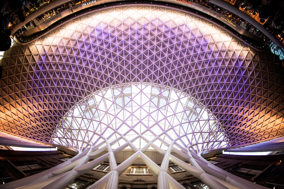 A large, modern indoor space featuring a domed ceiling with a geometric lattice pattern illuminated by purple and white lighting. The photograph is taken from a low angle, showcasing tall white structural columns supporting the ceiling. The environment appears spacious and well-lit, possibly within a commercial or public building, with natural light filtering through the lattice structure. In the background, parts of the interior walls and additional structural elements are visible, indicating an open-plan area suitable for events or large gatherings. This architectural scene, connected with home or commercial relocation logistics, highlights the scope and scale of modern building interiors where furniture transport and packing are involved during moves, fitting with services provided by Man With a Van King's Cross.