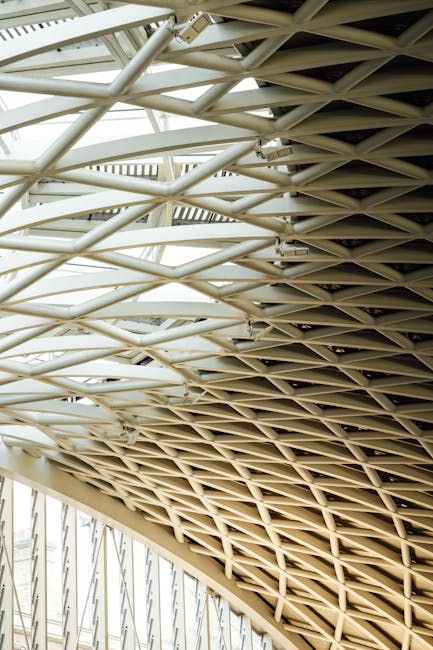 Close-up view of an intricate lattice roof structure made of beige and white metal beams arranged in a repeating geometric pattern, supporting a modern architectural roof with natural light filtering through. The design features intersecting horizontal and diagonal elements creating an open, airy ceiling. The structure appears to be part of a large interior space, possibly within a public building or a commercial property, with visible supports and glass panels framing the roof. This architectural detail emphasizes the use of structural steel components characteristic of contemporary building design, relevant to house removals and property relocations handled by Man With a Van King's Cross as part of their furniture transport and packing services during home relocation or moving projects.