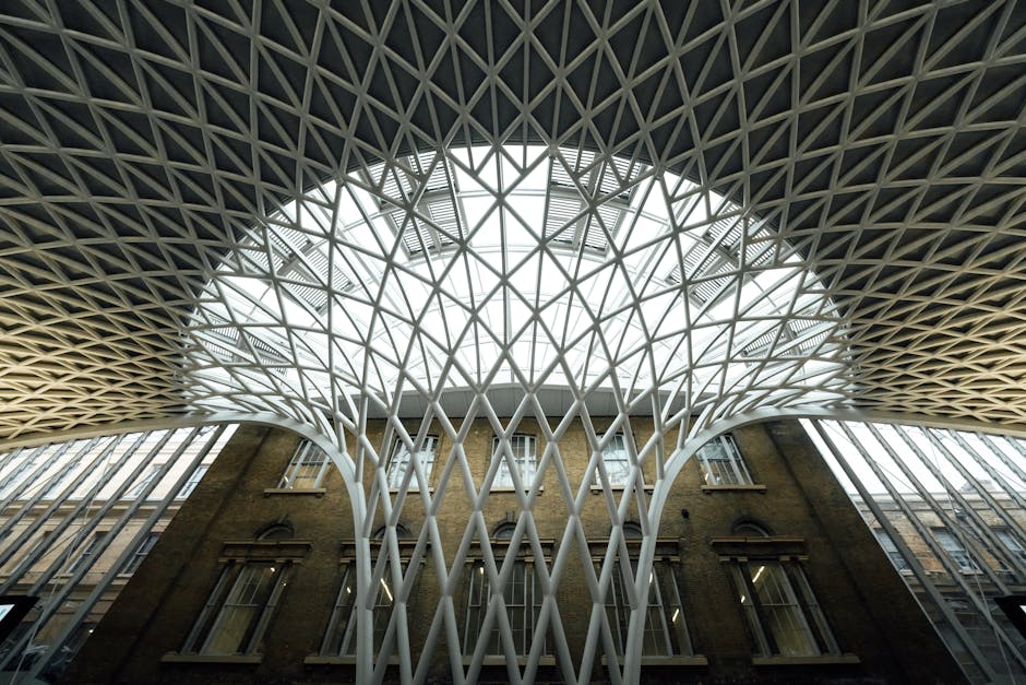 A large, modern indoor space featuring a domed ceiling with a geometric lattice pattern illuminated by purple and white lighting. The photograph is taken from a low angle, showcasing tall white structural columns supporting the ceiling. The environment appears spacious and well-lit, possibly within a commercial or public building, with natural light filtering through the lattice structure. In the background, parts of the interior walls and additional structural elements are visible, indicating an open-plan area suitable for events or large gatherings. This architectural scene, connected with home or commercial relocation logistics, highlights the scope and scale of modern building interiors where furniture transport and packing are involved during moves, fitting with services provided by Man With a Van King's Cross.