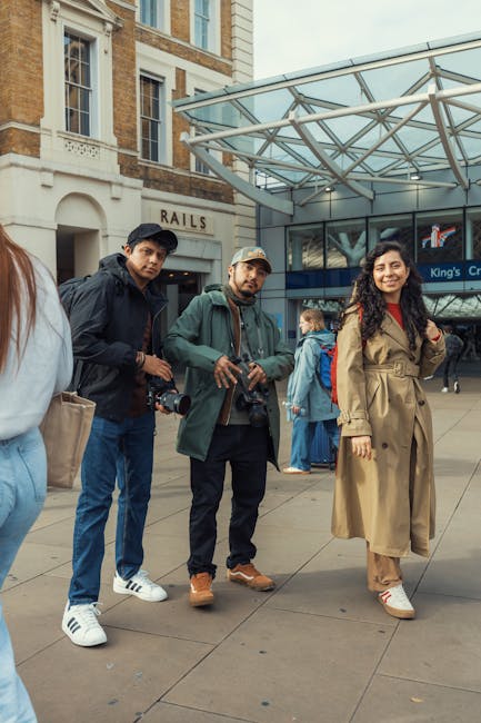 Outside Kings Cross station, three individuals stand on the pavement near the entrance with glass and metal awning overhead. The person on the left, wearing a white jacket and holding a beige bag, is partially visible. In the center, a man with a dark jacket, jeans, and white sneakers, holds a camera in his right hand and looks directly at the camera. Next to him, a man in a green jacket, beige cap, and brown shoes also holds a camera, with a neutral expression. To the right, a woman in a long beige trench coat, with dark curly hair, stands smiling, wearing white sneakers. The background shows a few other pedestrians, some with luggage, and a sign indicating 'King’s Cross' station entrance. This scene illustrates a typical busy day outside Kings Cross, relevant to house and furniture removals, as [COMPANY_NAME] may assist with packing and transportation logistics amid ongoing city regulations, including those discussed in the page about Camden Council rules affecting Kings Cross removals.