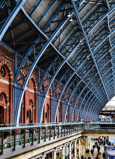 A spacious indoor area of a train station with a high, curved ceiling supported by blue steel arches and large glass panels, allowing natural light to illuminate the space. The lower level features a glass railing along a walkway with numerous people walking or standing, some near luggage or bags. Adjacent to the station's interior, visible red brick walls add a historic element to the architecture. In the background, a platform with trains and digital signage can be seen, indicating a busy transport hub. This setting reflects the typical environment for home relocation or furniture transport services, with the station interior ready to facilitate the movement of household items during a move, as seen in the context of house removals and logistics handled by companies like Man With a Van King's Cross.