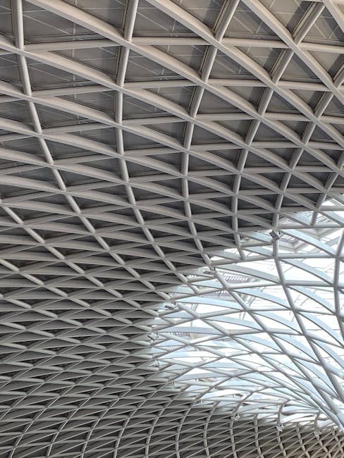 A detailed view of a large, modern architectural ceiling structure featuring a complex grid of metal beams and supports, with some sections incorporating glass panels that allow natural light to filter through. The steel framework is painted in a light grey color and forms an intricate geometric pattern, creating a spacious and airy environment. The ceiling design is part of a commercial or office building, which is consistent with the setting for house removals or office relocations. Although no furniture or moving equipment is visible in this image, the structural elements reflect the type of environment where professional removals such as those by Man With a Van King's Cross might occur, highlighting the importance of careful planning and logistics for furniture transport and packing within large, modern interiors.