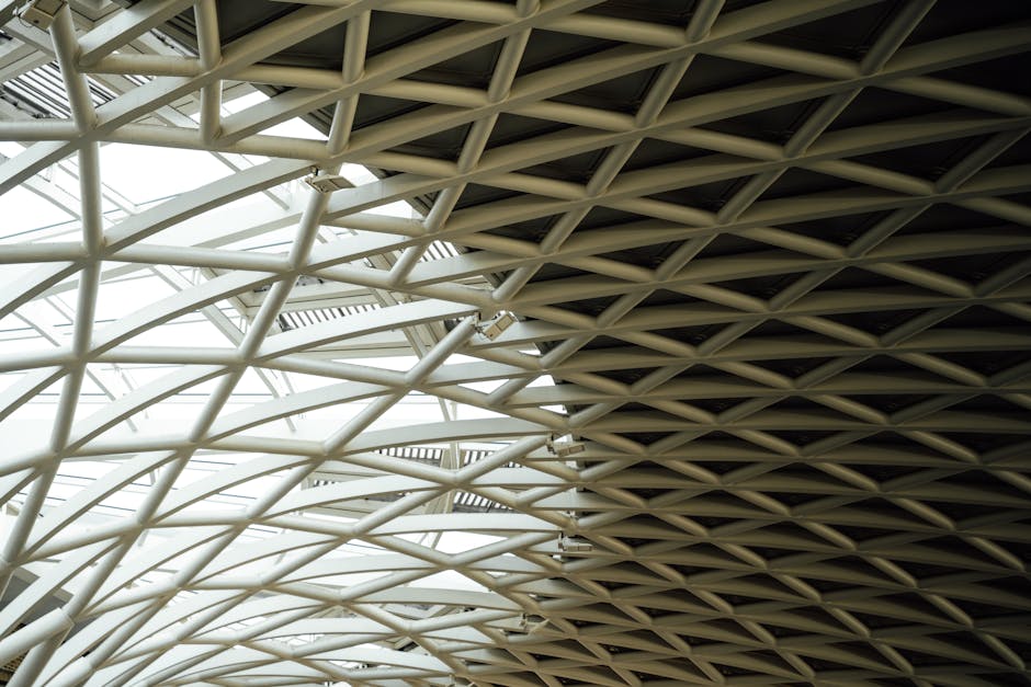 A detailed view of a modern architectural roof structure featuring a complex network of white metal beams and supports, with the left side illuminated by natural light from the skylights, while the right side appears darker with shadowed beams, illustrating the intricate design of a contemporary building's ceiling. The image captures the geometric pattern created by the intersecting beams, emphasizing the structural elements that could be part of a public or commercial space. This photograph is relevant to home relocation and moving services, as it depicts the environment where packing and loading processes may occur, aligning with the context of house removals and furniture transport managed by Man With a Van King's Cross. The focus on structural details underscores the importance of careful planning in loading and navigating such environments during a professional move.