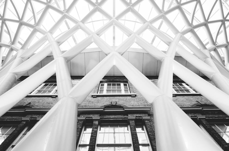 A black-and-white photograph taken from a low angle looking up at the exterior of a building, showcasing a modern architectural design with large, white steel cross-bracing structures forming a geometric pattern across the facade. The building features traditional brickwork with multiple rectangular windows framed in white, positioned between the steel supports. The sky is not visible due to the upward perspective, and the photograph highlights the contrast between the sleek steel framework and the brick elements. This structural design may be part of a contemporary office space or residential development in Kings Cross, with the image illustrating aspects of building construction or renovation associated with professional removals and relocation services from Man With a Van Kings Cross.