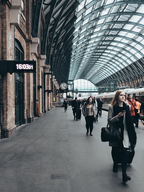 Inside a spacious train station with a high, arched glass and metal ceiling allowing natural light to fill the interior. The platform is populated with passengers, including a young woman in the foreground carrying a black handbag, wearing a light coat and holding a smartphone. Other travellers can be seen walking and standing near the platform, some dressed in casual clothing, with a few carrying luggage or backpacks. A digital departure board displaying the time and train information is mounted on a brick wall to the left, alongside large windows and an analog clock. In the background, a train is docked at the platform, visible through the glass walls, indicating a busy scene of travel and transit. This setting reflects a typical environment for home relocation and moving services, where collection and transfer of household items may occur, often involving packing supplies and moving equipment.