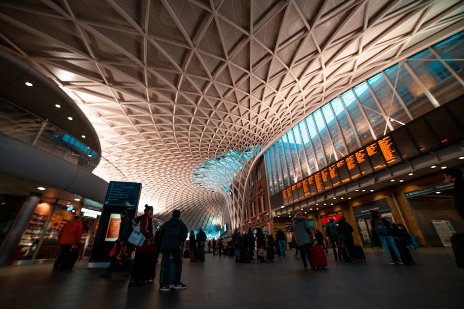 Inside a modern train station with a high, geometric patterned ceiling made of interconnected wooden or metal grid structures, illuminated by a combination of natural light from large glass windows and artificial lighting. Several passengers are waiting with luggage, including suitcases and carry bags, near digital departure boards displaying travel information in orange. Some individuals are standing or walking across the spacious, tiled floor, which features a mix of warm and cool lighting effects. In the background, retail outlets and service counters are visible along the perimeter. The scene captures a busy environment typical of a home relocation staging area where travel preparations are underway, with the potential for furniture and packing materials being transferred in or out. This setting reflects the logistical aspect of moving, emphasizing the importance of careful handling and planning in transportation or packing during home removals. Man With a Van King's Cross occasionally supports this process with professional furniture transport and packing services, as part of their house removals offering in Kings Cross.