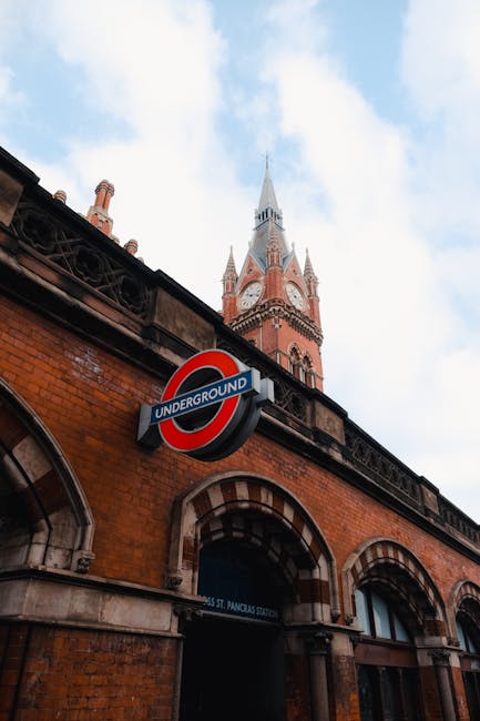A photograph taken during daylight hours shows a brick building with arched windows and a prominent round London Underground sign mounted on its façade, indicating an underground station entrance. Above the building, there is a tall, historic church tower with a pointed spire, constructed from red brick and grey stone accents, reaching into a partly cloudy sky. The scene provides context for urban house removals and relocating services, with the underground station sign suggesting nearby access for transportation logistics. The image captures the outdoor environment surrounding a characteristically historic London street, where Man With a Van King's Cross offers furniture transport and home relocation services, highlighting the area’s architecture and accessibility features relevant to moving operations.