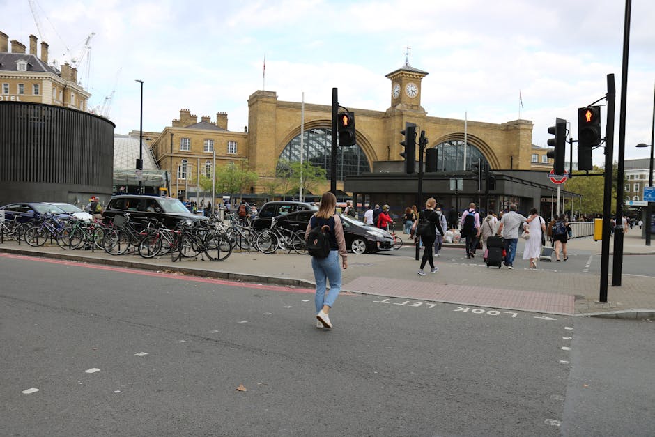 A busy scene outside King's Cross railway station in London featuring pedestrians crossing the road and walking along the pavement, some pulling wheeled luggage or carrying bags. On the left, there are several bicycles parked along a metal bike rack, and nearby, a black van is parked adjacent to a curb. The station's large, historic brick facade with a clock tower and arched windows is visible in the background under a cloudy sky. The area is illuminated by traffic lights, which are red for pedestrians. This setting is relevant to house removals and relocation logistics, with Man With a Van King's Cross providing moving services that involve parking and access considerations for home relocations, furniture transport, and packing and moving operations at busy transport hubs.